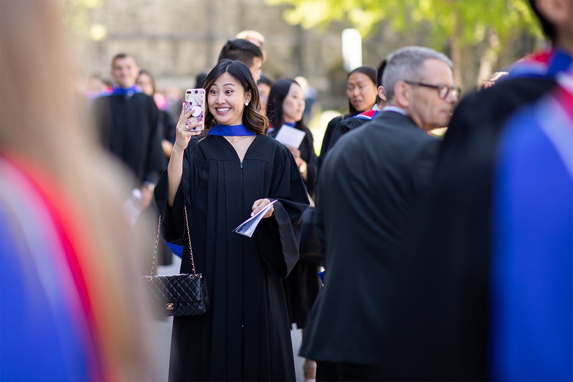 A woman in her graduation gown take a selfie just outside of convocation hall