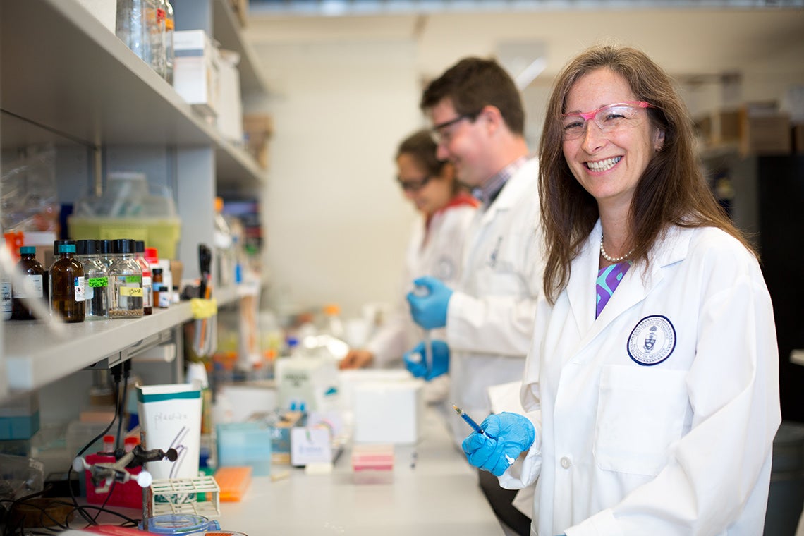 Molly Shoichet smiles for a photo in her lab. Lots of vials are visible and two students are working on the bench behind her