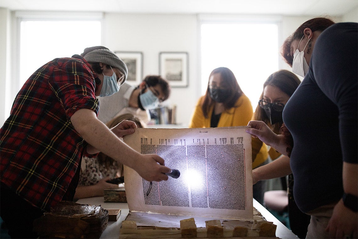 A student shines a flashlight through a page of a large book while Alexandra Gillespie examines the page from the opposite side