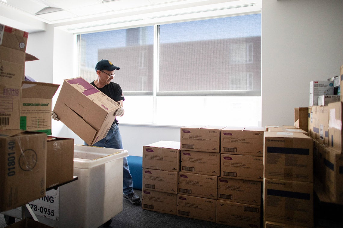 A worker lifts boxes of personal protective equipment supplies amidst stacks of other boxes