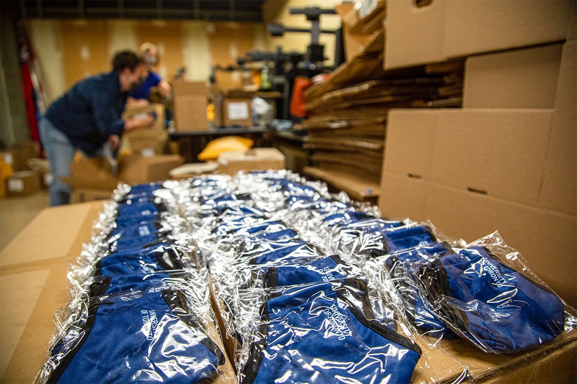 U of T masks laid out in a room with cardboard boxes. An employee unboxes the mask in the distance.