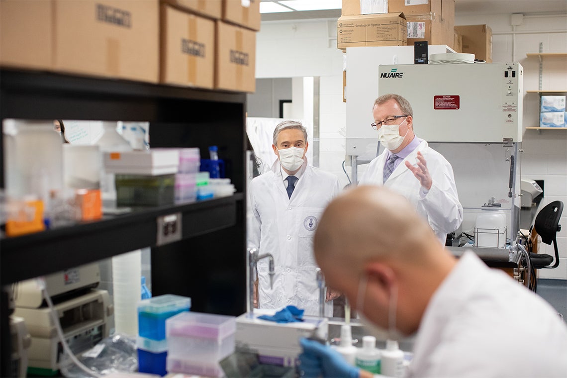 Scott Gray-Owen and François-Philippe Champagne wear masks and lab coats. They are watching a scientist work at a lab bench.