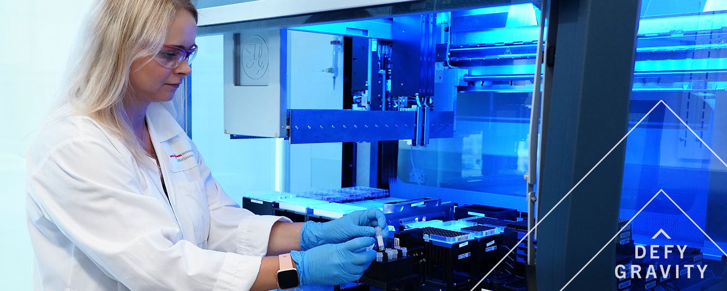A woman in a lab with the Defy Gravity wordmark