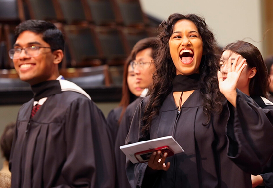 Students at convocation in robes smile and wave