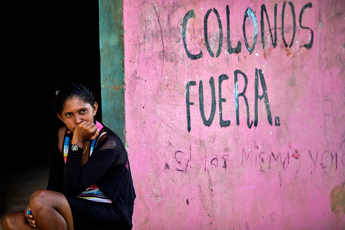 A Miskito woman sits next to a wall with graffiti saying "Settlers Out" 