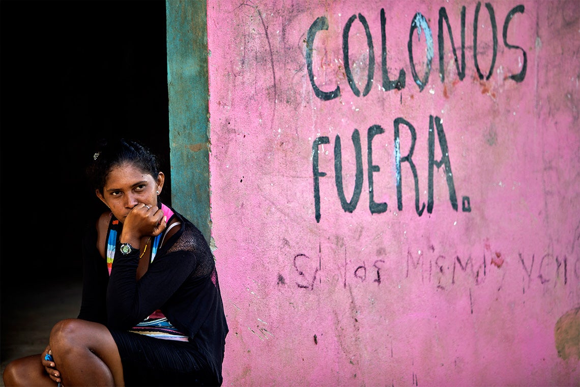 A Miskito woman sits next to a wall with graffiti saying "Settlers Out"