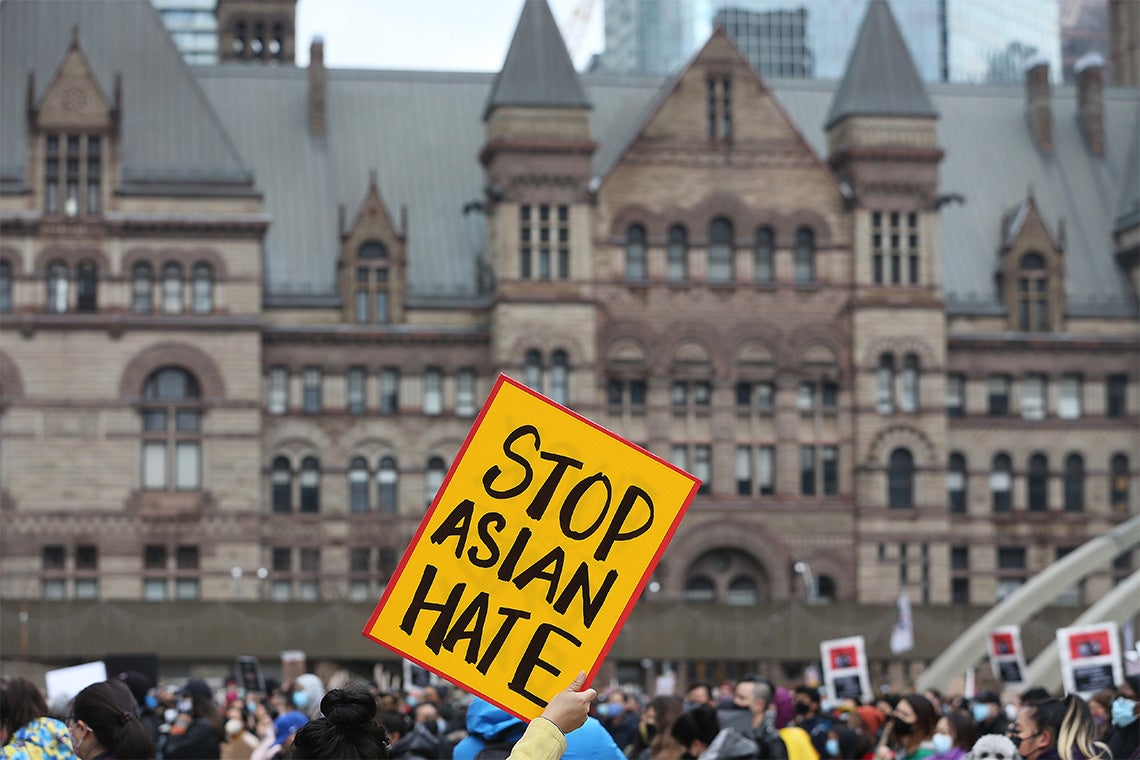 a person holds up a yellow poster that reads Stop Asian Hate. Queen's Park buildings are visible in the background