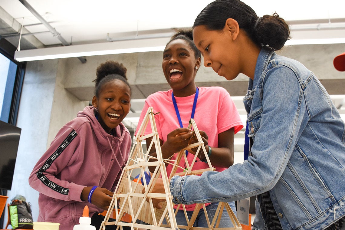 3 young women work on a structure made from popsicle sticks and glue while laughing