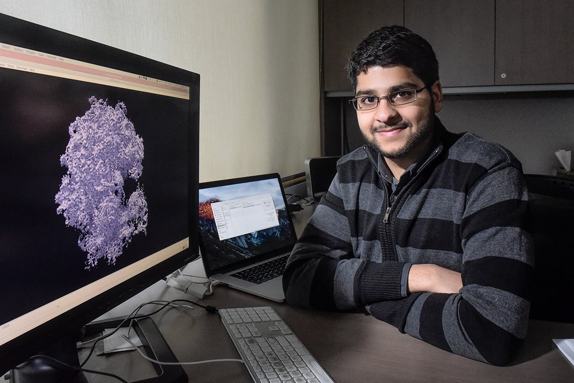 Ali Punjani sits in front of a computer screen displaying a 3D image of a protein molecule that was modelled using Structura Biotechnology’s cryoSPARC platform