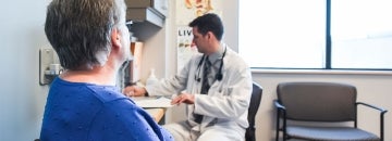 a doctor is seen writing while a senior citizen patient waits beside his desk