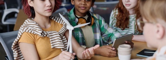 a young woman holds a model wind turbine. She is seated with other young people around a table.