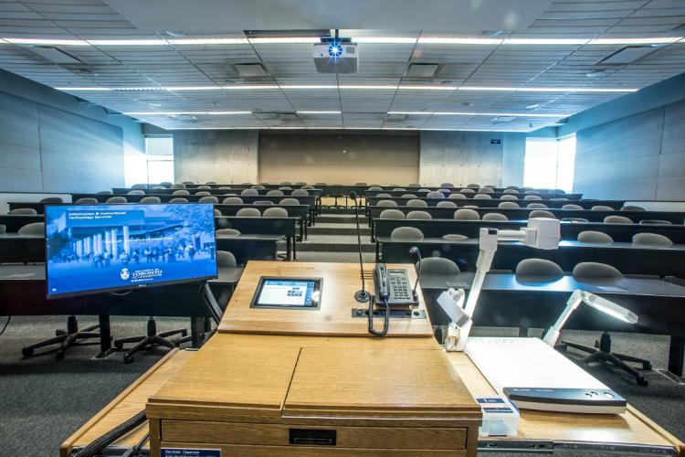 an empty classroom with a lectern in the foreground. There's a projector, monitor and computer at the lectern