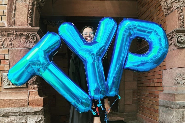 graduate holds up foil balloons that spell TYP