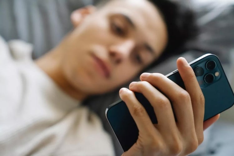 young man lies in bed looking at smartphone