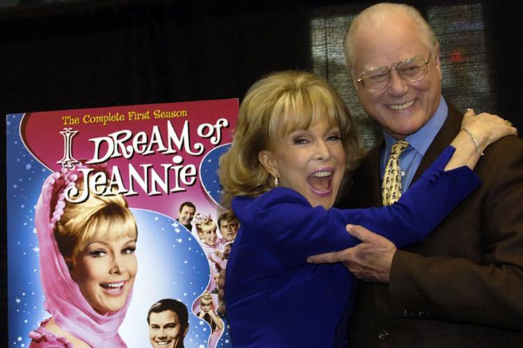‘I Dream of Jeannie’ co-stars Barbara Eden and Larry Hagman share an embrace in front of a 'I Dream of Jeannie' poster