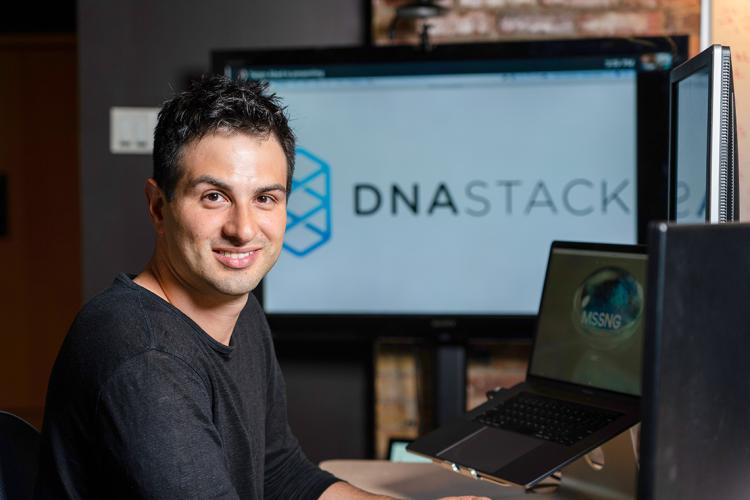 Marc Fiume sits at a computer, a monitor displaying the DNAstack logo is in the backbround
