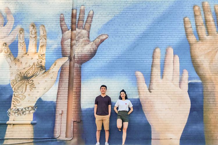 Weiwei Li and Catherine Chan stnad in front of a large mural on a wall depicting hands of different colours raised up