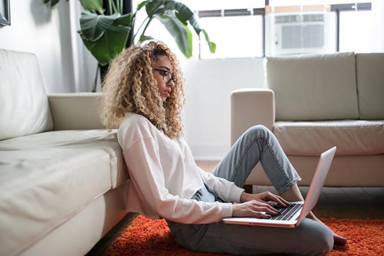 Woman sits on floor of apartment and works on laptop