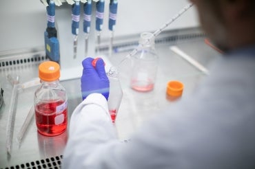 A researcher uses a pipette to drop fluid into a glass bottle behind a glass fume hood