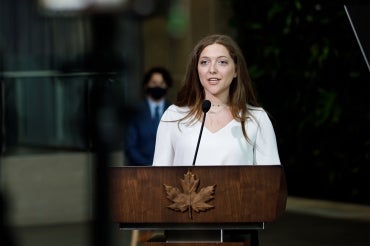 Lana El Sanyoura stands at a podium with Prime Minister Justin Trudeau visible in the distant background