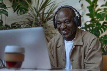An older man listens to music on earphones at a laptop while smiling