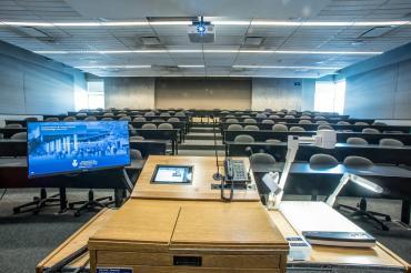 an empty classroom with a lectern in the foreground. There's a projector, monitor and computer at the lectern