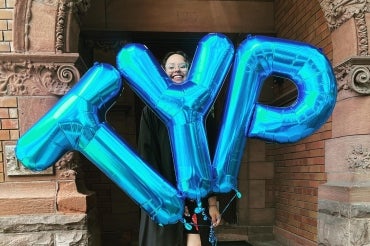 graduate holds up foil balloons that spell TYP