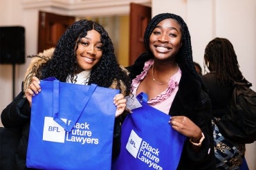Two smiling women hold up bright blue tote bags that say Black Future Lawyers