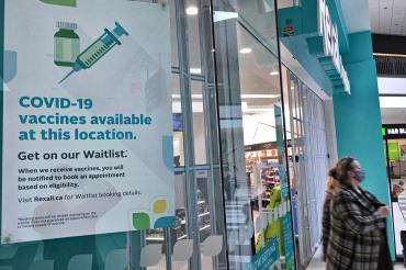 woman walks out of a Toronto pharmacy. Sign in forground reads "covid-10 vaccines available at this location"