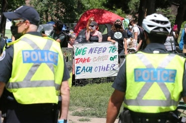 row of police in the foreground with protestors in the background. One of them holds a sign that says "we need to take care of each other"