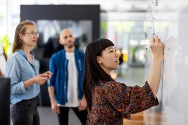 a young asian woman write on a whiteboard in an office while her colleagues look on