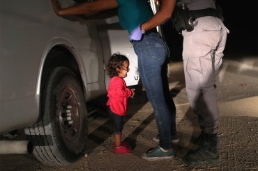 A young girl in a red sweater cries beside a white truck at night near the border while a border patrol officer detains her mother