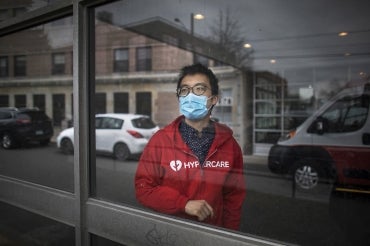 Albert Tai, wearing a blue mask and red 'Hypercare' hoodie, looks out a window at the street