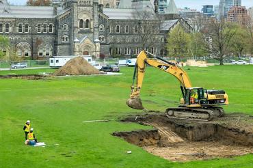 A backhoe digs a pit on Front Campus, with workers looking on and University College in the background