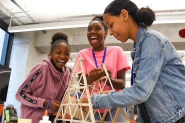 3 young women work on a structure made from popsicle sticks and glue while laughing