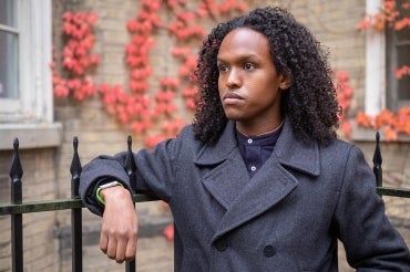 A photo of Egag Egag wearing a wool top coat leaning on a wrought iron fence with red ivy in the background