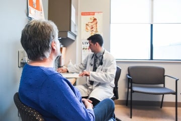 a doctor is seen writing while a senior citizen patient waits beside his desk