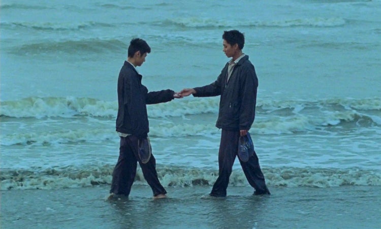 Main characters Nam and Việt touch hands standing on the shore of a beach