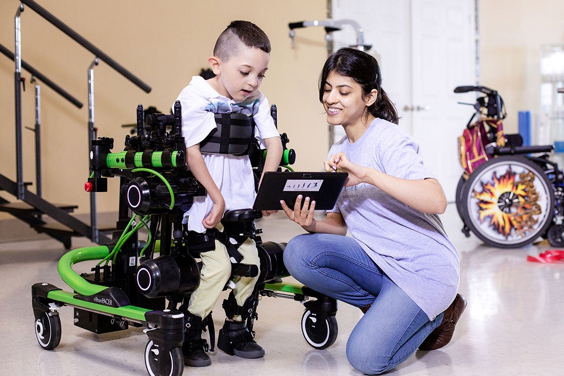 A small boy wearing Trexo Robotics assistive technology smiles as a helper shows him a tablet display